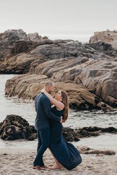 an engaged couple hugging on the beach in front of some rocks and water at sunset