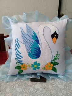 a white swan pillow with blue feathers and flowers on the front, sitting on a bed