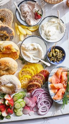 an assortment of food is laid out on a white tablecloth, including bagels, pickles, tomatoes, cucumbers and other foodstuffs