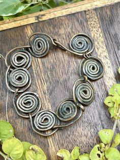 a bracelet made out of metal spirals sitting on top of a wooden table next to green leaves