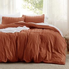 a bed with an orange comforter and two pillows in front of a white window