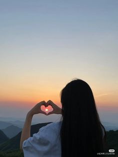 a woman making a heart shape with her hands at the top of a mountain during sunset