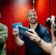 a man holding a blue book while standing next to another man in front of a red wall