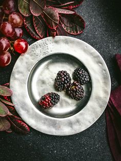 three raspberries on a silver plate next to some leaves and berries in the background