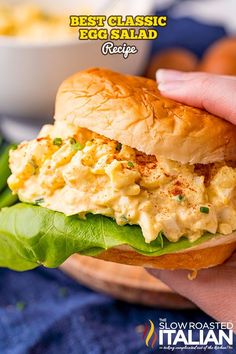 a hand holding a sandwich with egg salad on it in front of a bowl of macaroni and cheese