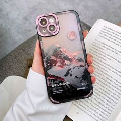 a person holding an iphone case with mountains on it and a book in their hand