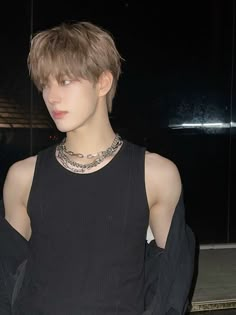 a young man with short hair wearing a black shirt and silver necklace standing in front of a window