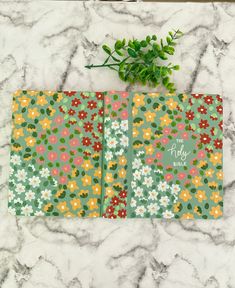 two notebooks with flowers on them sitting on a marble surface next to a plant