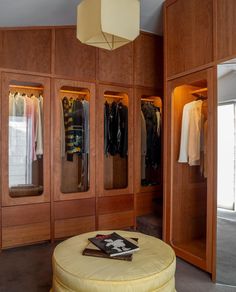 a room with wooden closets and clothes hanging on the walls, including a round ottoman