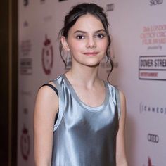 a young woman standing in front of a red carpet wearing a silver dress and earrings