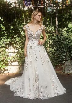 a woman standing in front of a fence wearing a wedding dress with flowers on it