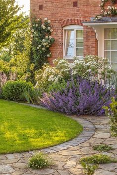 a brick house surrounded by lush green grass and purple flowers in the front garden area