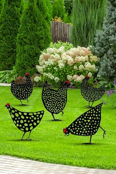 four metal chickens standing in the grass near some bushes and flowers on a sunny day