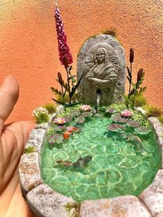 a hand is holding a miniature fountain with water and fish in it that has a buddha statue on top