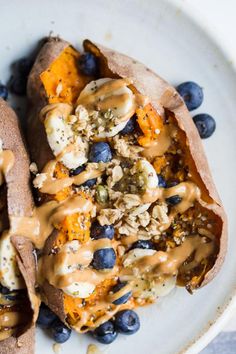 two stuffed sweet potatoes topped with blueberries and nuts on a white plate, covered in peanut butter