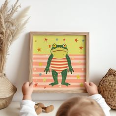 a young child holding up a framed frog art print in front of a white wall