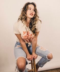 a woman sitting on top of a wooden stool wearing ripped jeans and a t - shirt