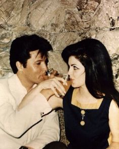 a man and woman sitting next to each other in front of a stone wall eating food