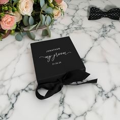 a black box with a bow tie and flowers in the background on a marble table