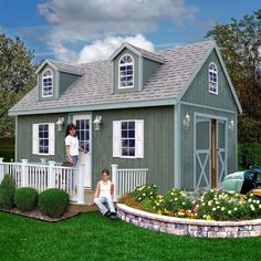 two people are standing in front of a small green house with white picket fence and flowers