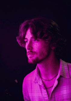 a man with long hair wearing a pink shirt and necklace in front of a black background