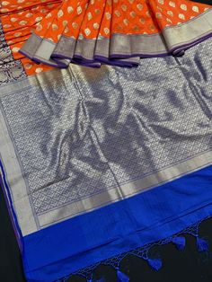 Traditional Orange Banarasi Silk Saree with Purple Borders and Blue Pallu with matching blouse piece. Saree is super soft!! Please note - Borders are wide. Approx. 7 1/2 inches Small Buttis all over the body of the Saree with Grand Pallu and Tassels Item : SareeColor : Orange/ Purple/Blue Base Fabric : Banarasi Silk SareeBlouse piece : Comes with Blouse pieceBlouse material : Banarasi Silk SareeFall & Edging done (Yes/No) : YesDisclaimer -:- Color variation is possible due to various reasons lik Designer Fusion Dupatta For Festivals, Fusion Style Designer Wear Dupatta For Festivals, Fusion Style Dupatta With Pallu For Navratri, Festive Fusion Saree With Pallu Detail, Festive Fusion Saree With Pallu, Fusion Style Traditional Wear With Pallu, Unstitched Fusion Traditional Wear For Diwali, Fusion Designer Traditional Wear For Festivals, Designer Fusion Traditional Wear For Festivals