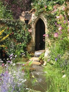 an outdoor garden with flowers and plants around the entrance to a stone building that is surrounded by greenery