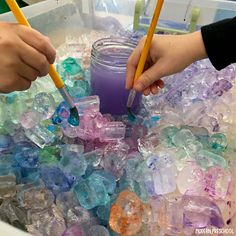 two hands are holding pencils in a container filled with ice cubes and purple liquid