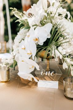 a table topped with white flowers and greenery next to a glass sign that says one