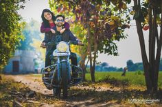 a man and woman riding on the back of a motorcycle down a dirt road next to trees