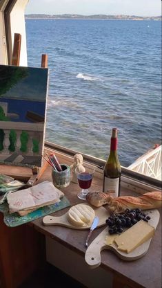 an easel with wine, cheese and bread on it next to a painting by the water