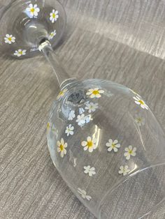 two wine glasses with white and yellow flowers on them sitting on a gray tablecloth