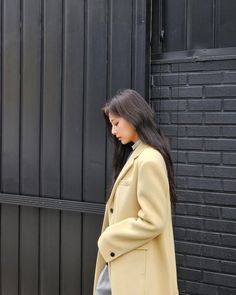 a woman standing in front of a black wall wearing a tan coat and grey pants