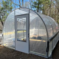 a small white greenhouse in the middle of some dirt and trees with a fence around it