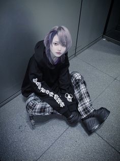 a person sitting on the ground wearing black and white plaid pants with purple hair, in front of a gray wall