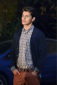 a young man standing in front of a car wearing a plaid shirt and blazer