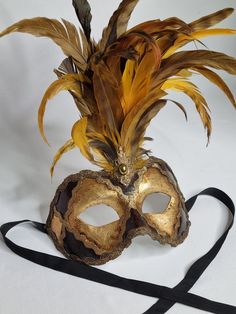 Elegant Venetian mask adorned with lush golden feathers, offering a refined and majestic look. Made according to the Venetian artisanal tradition, this mask is perfect for masked balls, carnivals and costume parties. It features detailed gold hardware and a central embellishment with a gold-tone costume jewel, adding a touch of sophistication to the overall look. Traditional Gold Masks For Carnival, Traditional Masks For Mardi Gras Carnival, Gold Costume Mask For Mardi Gras, Gold Masks For Costume Festivals, Traditional Masks And Prosthetics For Mardi Gras Carnival, Gold Costume Masks For Festivals, Traditional Gold Masks And Prosthetics For Carnival, Gold Mardi Gras Costume Mask, Traditional Gold Masquerade Mask For Carnival