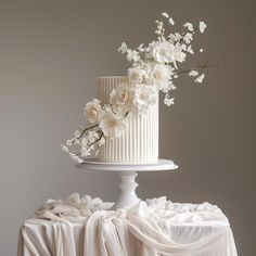 a white wedding cake with flowers on the top is sitting on a draped tablecloth