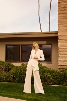 a woman standing in front of a house with her hands on her hips and wearing a white suit