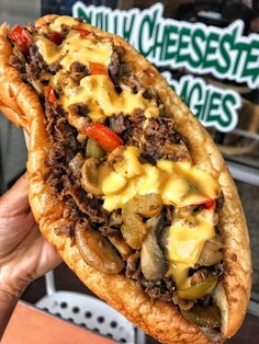 a person holding up a sandwich with cheese and meat on it in front of a sign