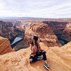 a woman sitting on the edge of a cliff looking out over canyons and river