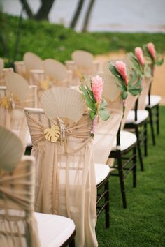 rows of white folding chairs with pink flowers and green leaves tied to them on the grass