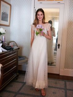 a woman in a white dress is standing by a door and holding some pink flowers