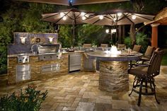 an outdoor kitchen is lit up at night with umbrellas over the grill and table