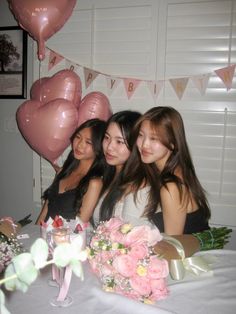 three young women sitting at a table with pink balloons and flowers in front of them