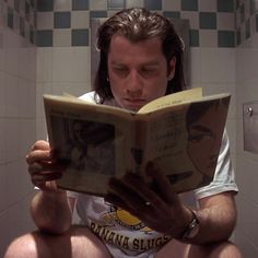 a man reading a book while sitting on the toilet in a bathroom with tiled walls