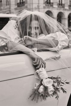 a bride leaning on the back of a car with her veil over her head and flowers in her hand