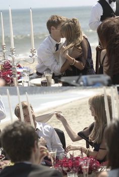 people sitting at a table on the beach with candles and flowers in front of them