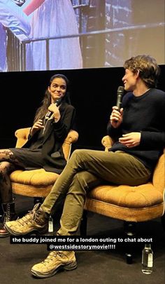 two people sitting in chairs talking to each other with a screen on the wall behind them