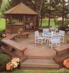 a wooden deck with white chairs and table next to flowers on the ground in front of a gazebo